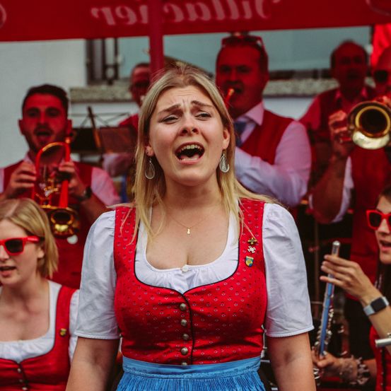 Eine blonde Frau in einem traditionellen roten Kleid singt, während sie vor einer Blechbläserband unter einem roten Schirm steht.