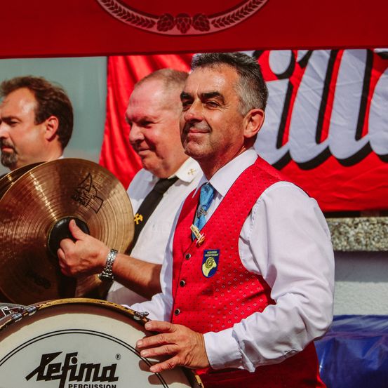 Ein Mann in einem roten Weste mit Logo lächelt und hält eine Trommel und Becken. Hinter ihm hält ein anderer Mann ein Becken.