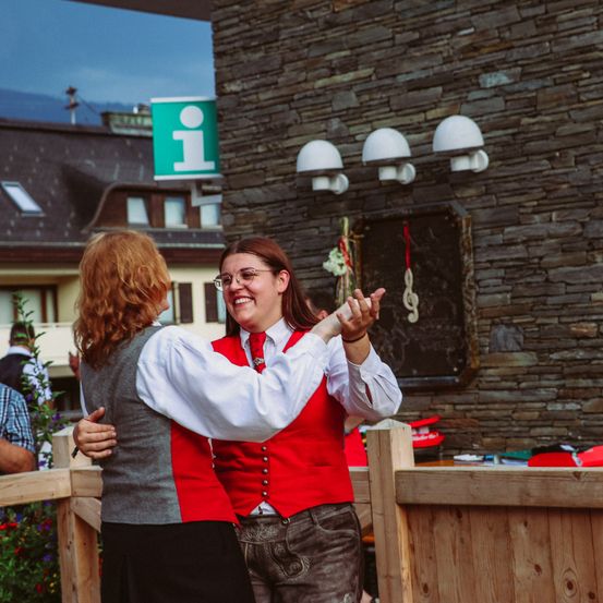 Zwei Frauen tanzen vor einem Steingebäude, eine trägt ein rotes Weste und die andere eine graue Weste. Sie sind von einem Holzzaun umgeben, mit einem Schild und einem Haus im Hintergrund.