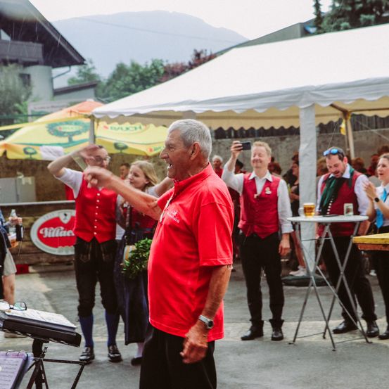 Eine Gruppe von Menschen steht draußen auf einem Festival. Ein älterer Mann in einem roten Shirt lächelt und schaut zur Kamera. Eine Frau mit einem Kind ist ebenfalls im Bild, und ein Tisch mit Getränken ist sichtbar.