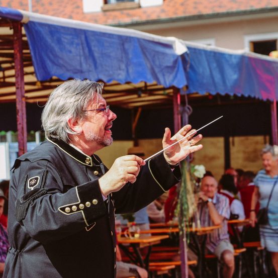 Ein älterer Mann mit grauem Haar, der eine Brille und eine schwarze Uniform trägt, dirigiert eine Band unter einem blauen Vordach bei einer Outdoor-Veranstaltung. Hinter ihm sitzen Leute an Tischen.