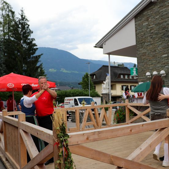 Ein Paar tanzt auf einer Holzterrasse mit einem Holzzaun, umgeben von einer Bergkulisse und einem roten Schirm.