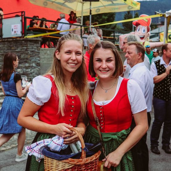 Zwei Mädchen in traditioneller bayerischer Kleidung, lächelnd und posierend für ein Foto. Ein Korb und eine Tischdecke sind in ihren Händen. Dahinter stehen andere Leute und eine große Comicfigur.