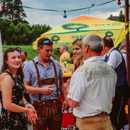 Ein Mann und eine Frau in traditioneller bayerischer Kleidung unterhalten sich mit einer Gruppe von Menschen unter einem gelben Zelt, während andere herumstehen. Im Hintergrund ist ein verschwommenes Bild eines Mannes, einer Frau und eines Zeltes zu sehen. Der Himmel ist bewölkt und es sind einige Bäume in der Ferne.