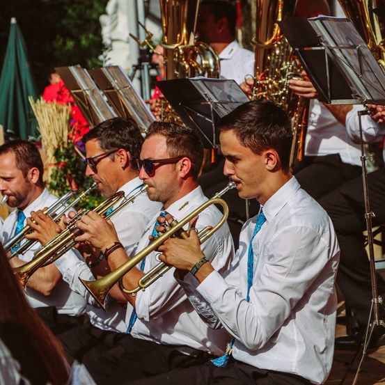 Eine Gruppe von Musikern in weißen Hemden und blauen Krawatten spielt Trompeten auf einer Freiluftbühne. Hinter ihnen spielen andere Posaunen und Tuben.