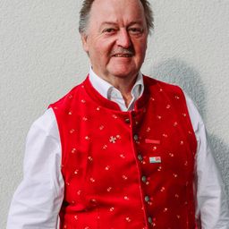 Ein lächelnder Mann in einem rot bestickten Weste mit einem weißen Hemd und weißem Unterhemd posiert vor einer weißen Wand.