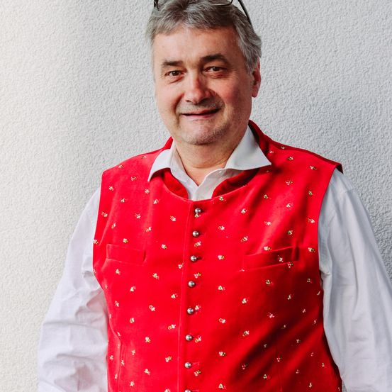 Ein Mann mit einer roten, bestickten Weste über einem weißen Hemd, steht vor einer weißen Wand und lächelt.