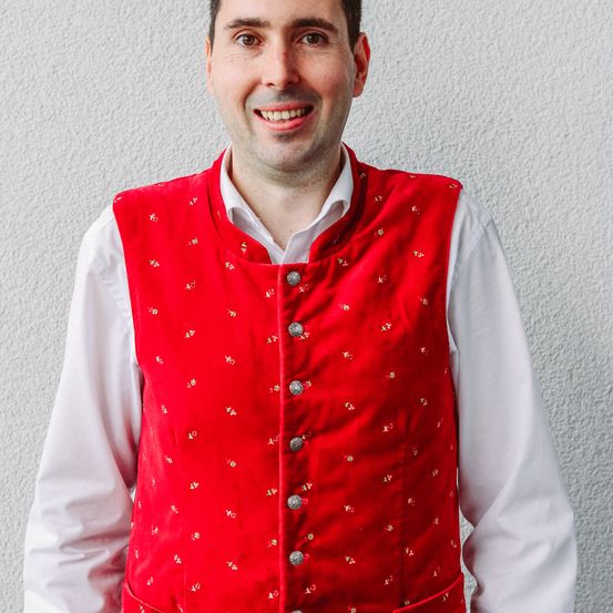 Ein Mann in einer roten Weste mit goldener Stickerei und silbernen Knöpfen lächelt und steht vor einer weißen Wand.