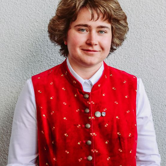 Eine Person in einem roten Weste mit Blumenmuster und einem weißen Hemd, die gegen eine weiße Wand steht und für ein Foto lächelt.