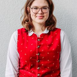 Eine junge Frau in einem roten Weste mit goldenen Blumenmustern und einem weißen Hemd lächelt und posiert für ein Foto.