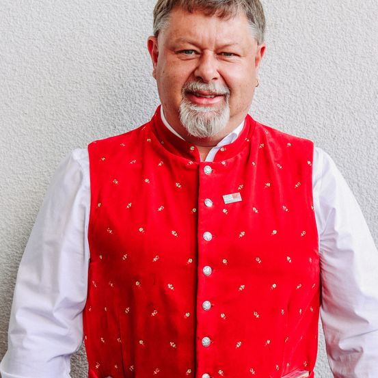 Ein Mann in einer roten Weste mit weißem Innenshirt steht vor einer weißen Wand.