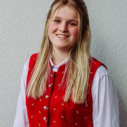 Eine lächelnde Frau mit blonden Haaren, die eine rot bestickte Weste über einem weißen Langarmhemd trägt und gegen eine weiße Wand steht.