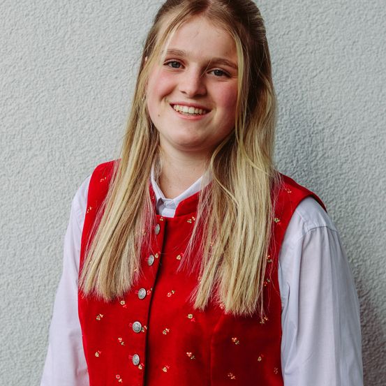 Eine lächelnde Frau mit blonden Haaren, die eine rot bestickte Weste über einem weißen Langarmhemd trägt und gegen eine weiße Wand steht.