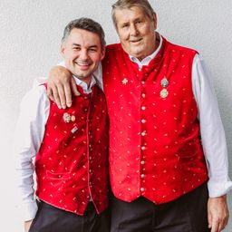 Zwei Männer, die rote Westen mit dekorativen Mustern tragen, posieren gemeinsam vor einer weißen Wand. Der ältere Mann hat seine Hand auf der Schulter des jüngeren Mannes.