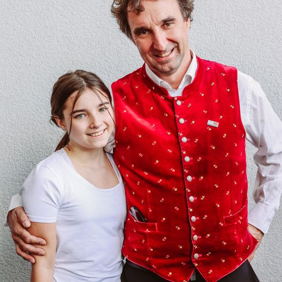 Ein Mann in einer roten Weste und ein Mädchen in einem weißen T-Shirt posieren für ein Foto. Der Mann lächelt und hat seine Hand auf der Schulter des Mädchens. Das Mädchen lächelt und hat seine Hand auf der Schulter des Mannes. Der Mann hat eine Anstecknadel auf seiner Weste.