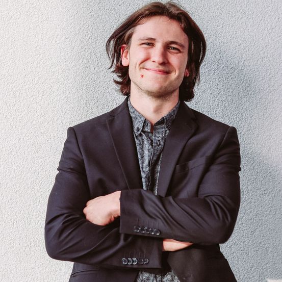 Ein Mann mit langen, welligen Haaren lächelt und posiert für ein Foto vor einer weißen Wand, trägt eine schwarze Anzugjacke und ein gemustertes Hemd.