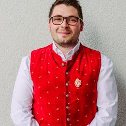 Ein Mann mit Brille steht gegen eine Wand und trägt eine rote Weste mit Blumenbrosche.