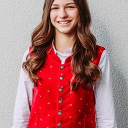 Ein junges Mädchen mit langen braunen Haaren, das eine rot bestickte Weste und ein weißes Hemd trägt, lächelt und posiert für ein Foto vor einer weißen Wand.