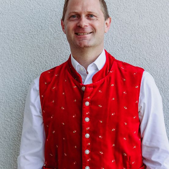 Ein Mann steht vor einer weißen Wand und lächelt. Er trägt einen roten Weste mit goldenen Knöpfen und ein weißes Hemd.