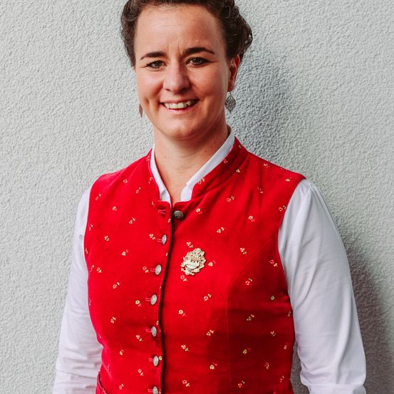 Nahaufnahme einer lächelnden Frau in einer rot bestickten Weste über einem weißen Langarmshirt.