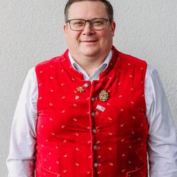 Ein Mann steht vor einer weißen Wand. Er trägt eine rote Weste mit Goldstickerei, ein weißes Langarmhemd und eine Brille.