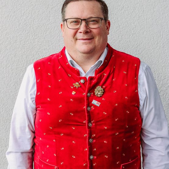 Ein Mann steht vor einer weißen Wand. Er trägt eine rote Weste mit Goldstickerei, ein weißes Langarmhemd und eine Brille.