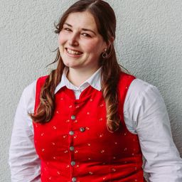 Eine lächelnde Frau in einem roten Weste mit dekorativen Mustern und einem weißen Hemd steht vor einer weißen Wand.