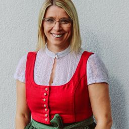 Eine Frau mit blondem Haar steht vor einer weißen Wand, lächelt und trägt eine Brille, ein rotes Westchen und eine weiße Spitzenbluse.