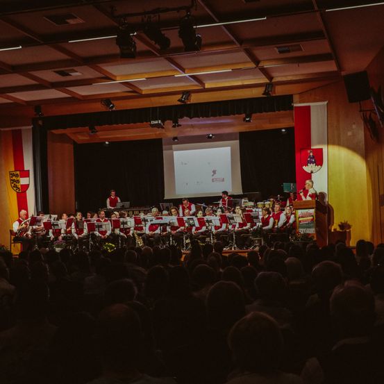 Ein großes Publikum sitzt vor einer Bühne, auf der eine Band auftritt. Hinter der Band befindet sich ein Podium mit Mikrofon und eine große Leinwand.
