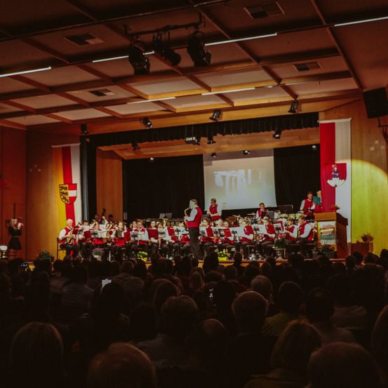 Ein Konzert einer Blaskapelle findet in einer großen Halle statt, mit einer Projektionsleinwand und Flaggen auf der Bühne.