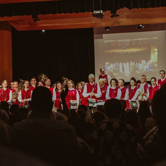 Ein großes Publikum beobachtet eine musikalische Aufführung auf der Bühne, bei der Musiker in roten Westen verschiedene Instrumente spielen, begleitet von einer großen Leinwand dahinter.