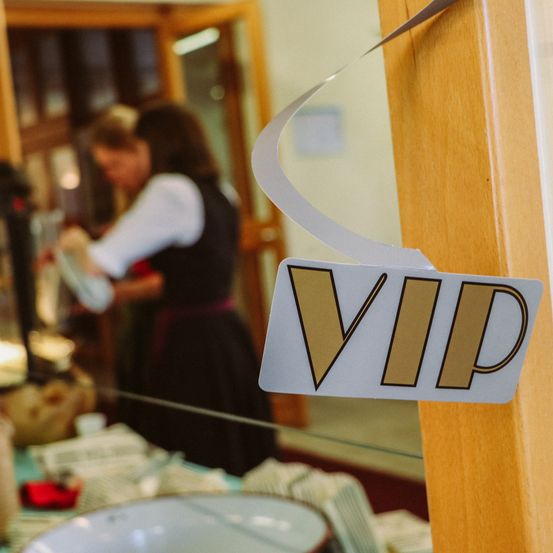 Ein VIP-Schild hängt an einer Holztür. Dahinter steht eine Frau in einem verschwommenen Hintergrund, möglicherweise ist sie dabei, etwas zuzubereiten.