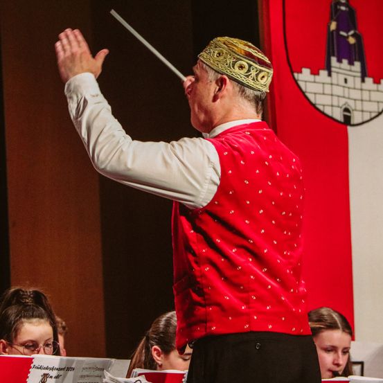 Ein älterer Mann mit einem roten Vest und einem goldenen Hut hält einen Taktstock und dirigiert vermutlich ein Orchester. Die Orchestermitglieder sitzen und schauen ihn an. An der Wand hinter ihnen hängt ein roter Banner mit einer Burg und einer Frau darauf.