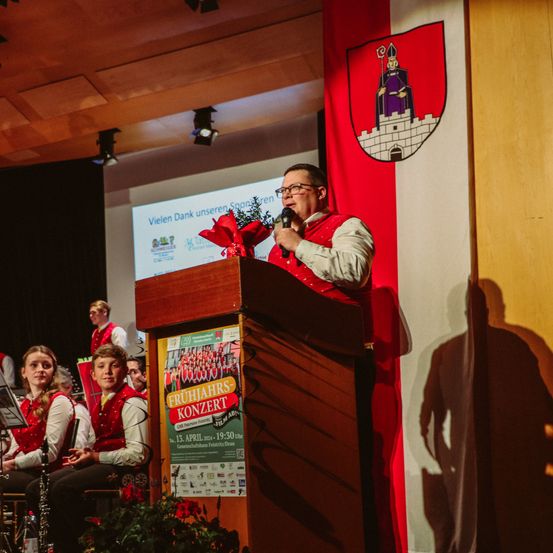 Ein Mann in Rot spricht in ein Mikrofon an einem Rednerpult mit einem Schild davor. Dahinter befindet sich ein rot-weißes Banner mit einem Burglogo.