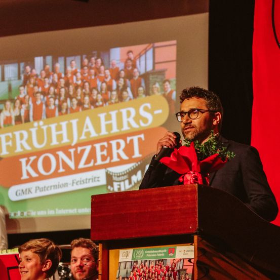 Ein Mann in einem Anzug steht an einem Rednerpult mit einem Mikrofon und einem Blumenstrauß vor einer Projektionsleinwand, auf der 'Frühjahrs-Konzert' steht.