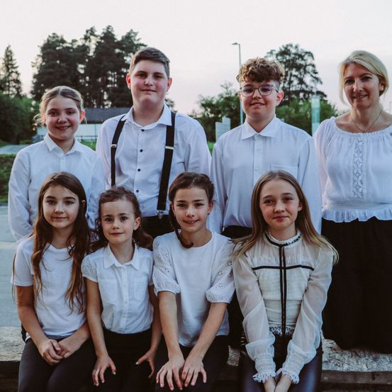 Eine Gruppe von Kindern in weißen Hemden und einer Frau in einer weißen Bluse posiert für ein Foto auf einer Bank vor Bäumen.