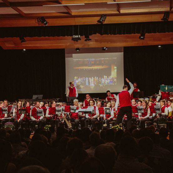 Ein Orchester spielt auf der Bühne in einem großen Saal, während eine Menge zuschaut. Ein Mann in Rot steht auf der rechten Seite.