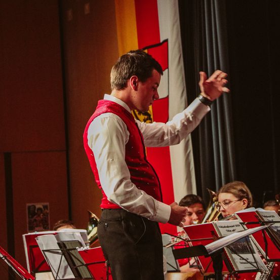 Ein Dirigent in einem roten Vest dirigiert ein Orchester. Die Musiker sitzen und spielen verschiedene Instrumente, darunter Trompete und Posaune. Im Hintergrund ist eine rot-weiße Flagge zu sehen.