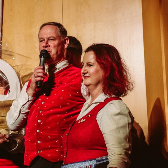 Ein Mann und eine Frau in roten traditionellen Kostümen, der Mann hält ein Mikrofon und die Frau lächelt, während sie dicht nebeneinander vor einer Holzwand stehen.