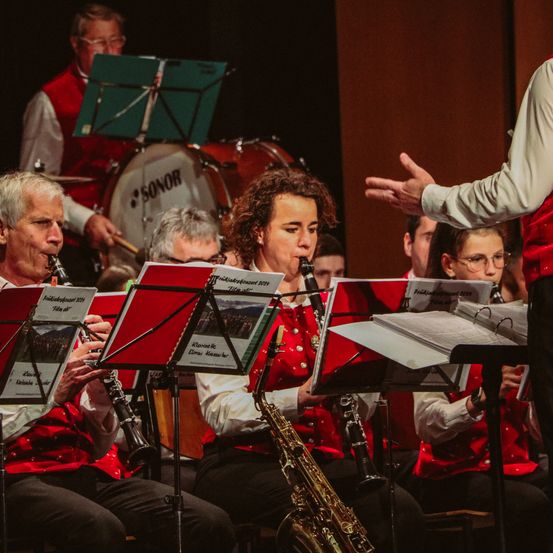 Eine Band von Musikern spielt auf der Bühne. Der Dirigent steht vor ihnen. Die Musiker sitzen auf Stühlen und spielen ihre Instrumente, darunter Klarinetten, Saxophone und Trommeln.