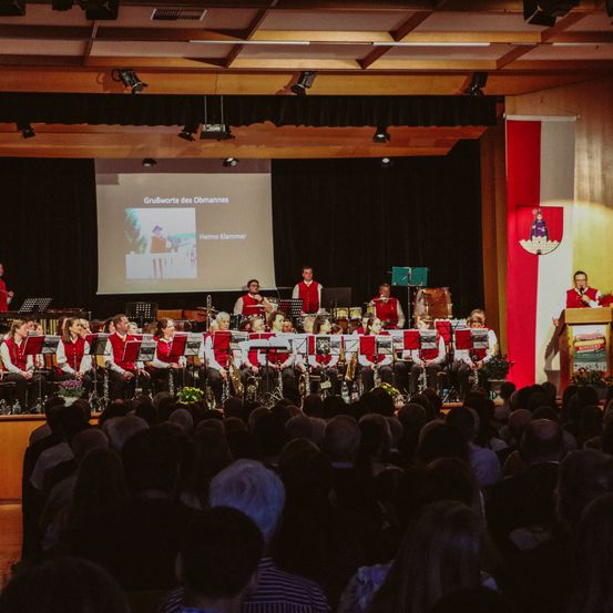 Eine Gruppe von Musikern tritt auf der Bühne vor einem Publikum auf. Eine Projektionsleinwand zeigt ein Bild eines Mannes.