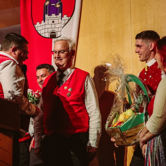 Ein älterer Mann in einem roten Weste wird bei einer Zeremonie mit einem Obstkorb ausgezeichnet.