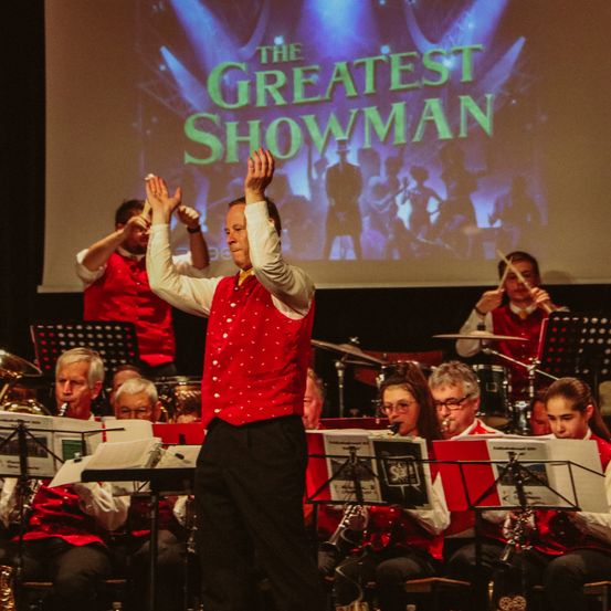 Ein Mann in einem roten Weste tritt mit einer Band auf der Bühne auf. Sie spielen verschiedene Instrumente, und hinter ihnen befindet sich eine Projektionsleinwand mit den Worten 'The Greatest Showman'.