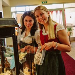 Zwei Frauen, die lächeln und traditionelle Kleidung tragen, stehen vor einer Popcornmaschine, eine von ihnen hält eine Popcorntüte.