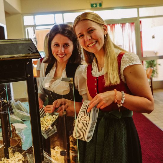 Zwei Frauen, die lächeln und traditionelle Kleidung tragen, stehen vor einer Popcornmaschine, eine von ihnen hält eine Popcorntüte.