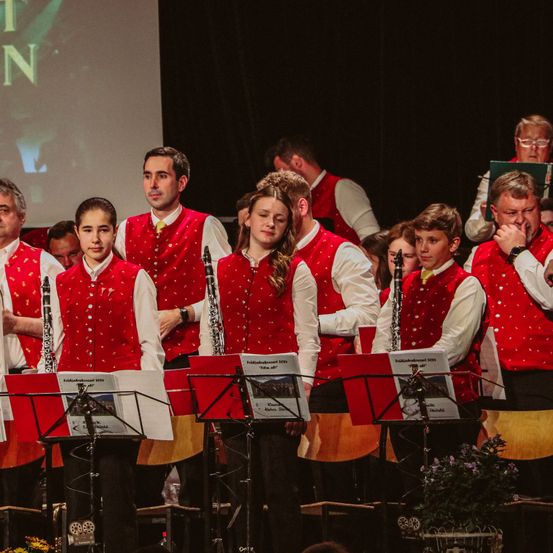 Ein Chor junger Musiker in roten Westen mit weißen Punkten tritt mit Notenblättern, Klarinetten und einer Projektionsleinwand im Hintergrund auf der Bühne auf.