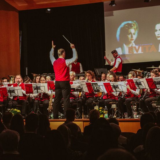 Ein Orchester tritt auf der Bühne auf, und der Dirigent hebt seinen Taktstock in die Luft. Die Musiker spielen ihre Instrumente, während eine große Leinwand hinter ihnen den Film "Evita" zeigt.