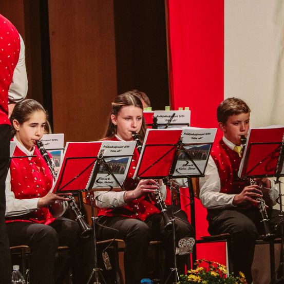 Drei Kinder in roten Westen spielen Klarinette, während sie bei einer musikalischen Aufführung auf Stühlen sitzen.