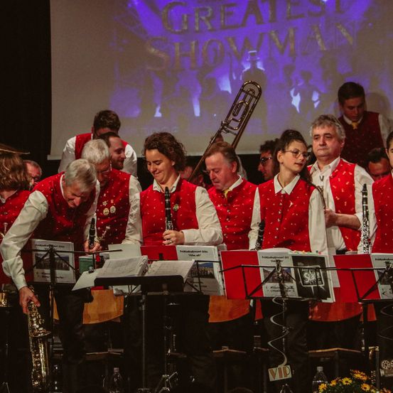 Ein Blasorchester steht auf der Bühne, mit einer Projektionsleinwand hinter ihnen, auf der 'Greatest Showman' steht. Die Musiker sind in roten und weißen Westen gekleidet und halten ihre Instrumente.