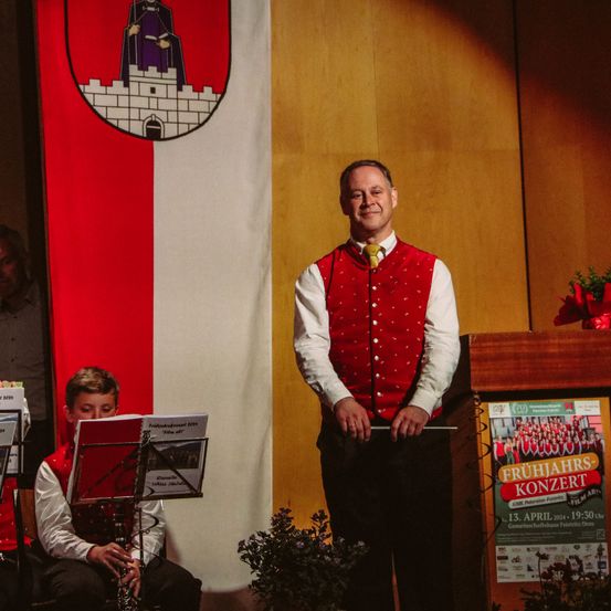 Ein Mann mit rotem Weste steht auf der Bühne und hält einen Taktstock vor einem Podium mit Frühlingsarrangement. Hinter ihm spielt ein junger Junge Klarinette. Eine Flagge mit Wappen und einer Person im Hintergrund.